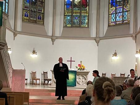 Innenraum der bunt beleuchteten Evangelischen Stadtkirche in Baden-Baden während des Aperol-Gottesdienstes am 13. September 2025. Die Besucher sitzen und schauen nach vorn, wo Dekan Christian Link in seiner schwarzen Robe vor dem Altar steht und seine Predigt hält.